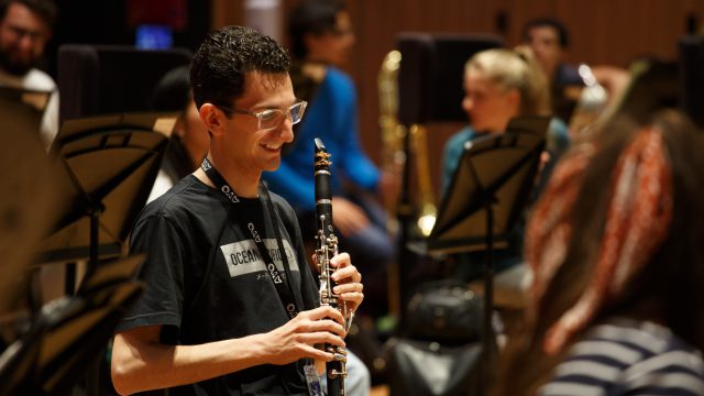 AYO musician pictured holding a clarinet and smiling. He is wearing glasses and an AYO t-shirt.