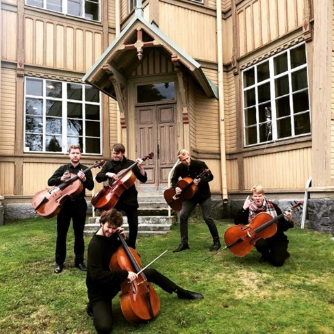 Five musicians dressed in black posing with cellos, on the lawn outside a wooden building.