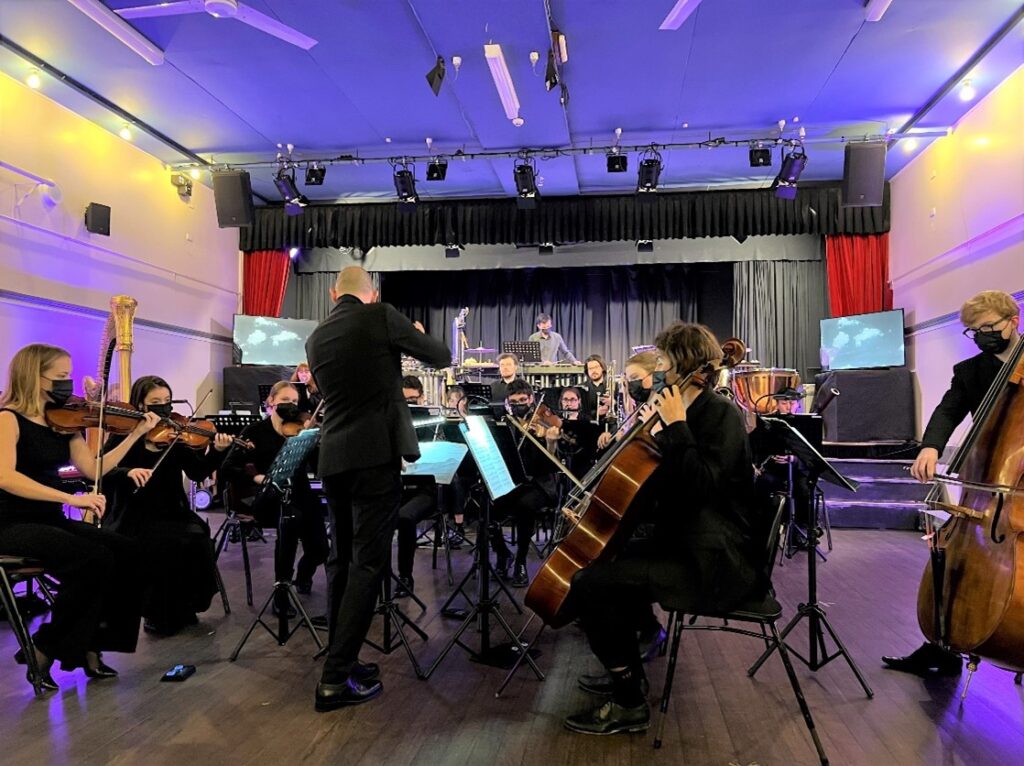 A small ensemble of musicians wearing concert blacks perform on stage, guided by a conductor. The room is lit by a purple wash of light.