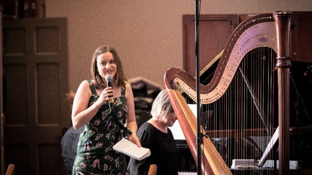 AYO Composition participant Sophie Van Dijk speaks into a microphone. She is smiling and standing next to a harp.