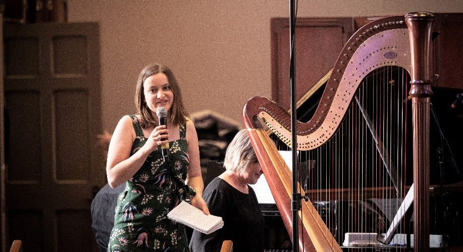 AYO Composition participant Sophie Van Dijk speaks into a microphone. She is smiling and standing next to a harp.