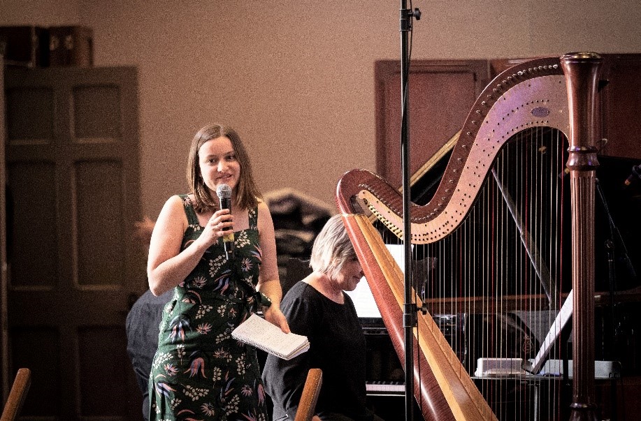 AYO Composition participant Sophie Van Dijk speaks into a microphone. She is smiling and standing next to a harp.