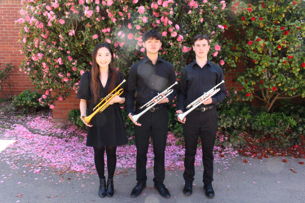 Three trumpet players stand in front of a flowering bush. They are dressed in concert blacks and are smiling at the camera while holding their instruments.