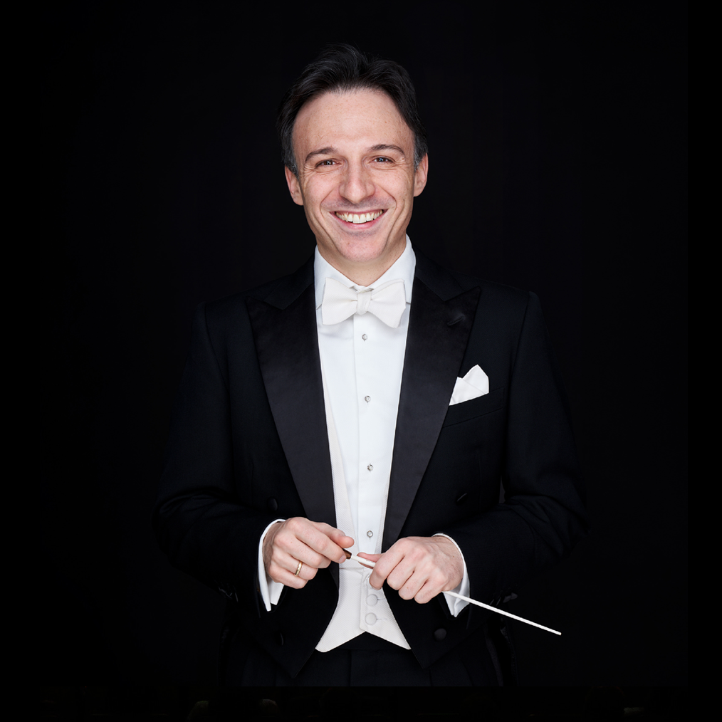 Conductor Umberto Clerici smiles at the camera while holding a conductor's baton in both hands. He is wearing a black suit.