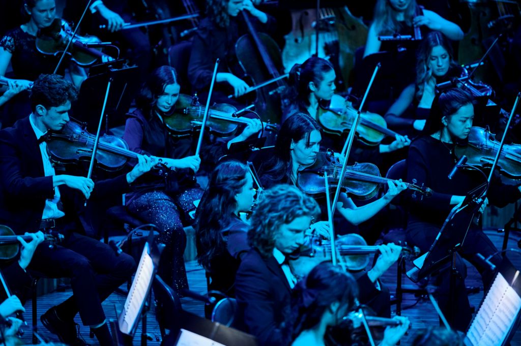 An orchestra of young musicians perform on stage, lit in blue light.