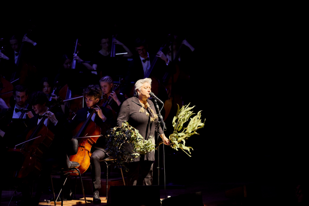 Dr Lou Bennett AM performs on stage while holding branches of gum leaves in each hand.