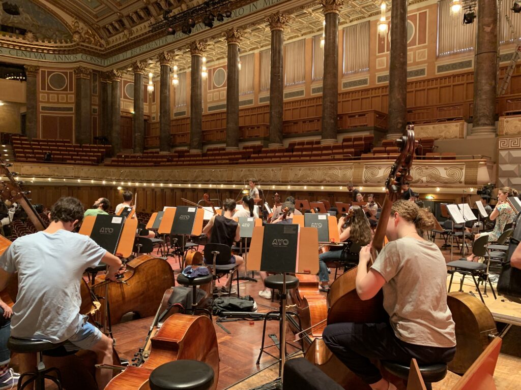 An orchestra rehearses in a concert hall with ornate chandeliers hanging from the ceiling.