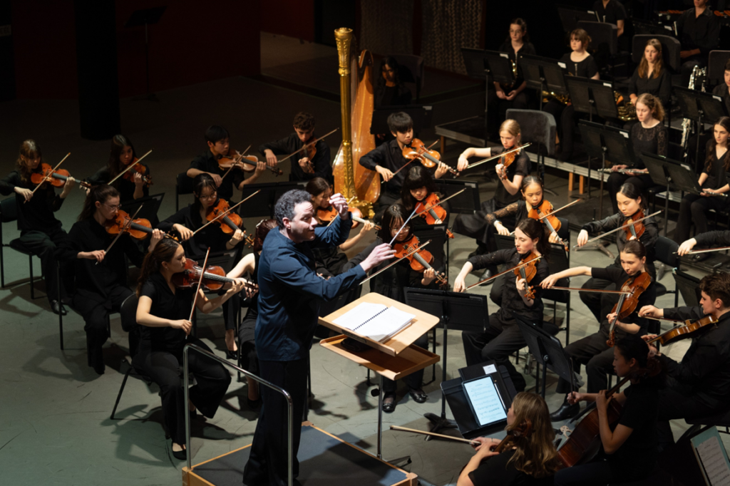 An orchestra of young musicians in concert blacks perform on stage.