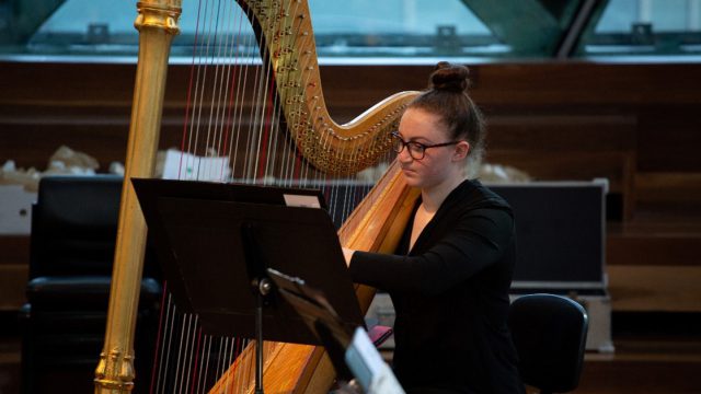 A young woman with dark brown hair in a bun plays a large golden harp on stage.