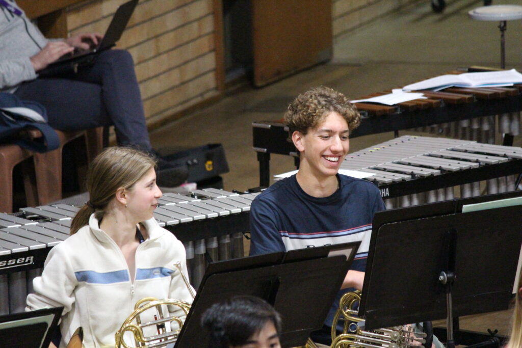 Two French Horn players smile during an orchestra rehearsal.