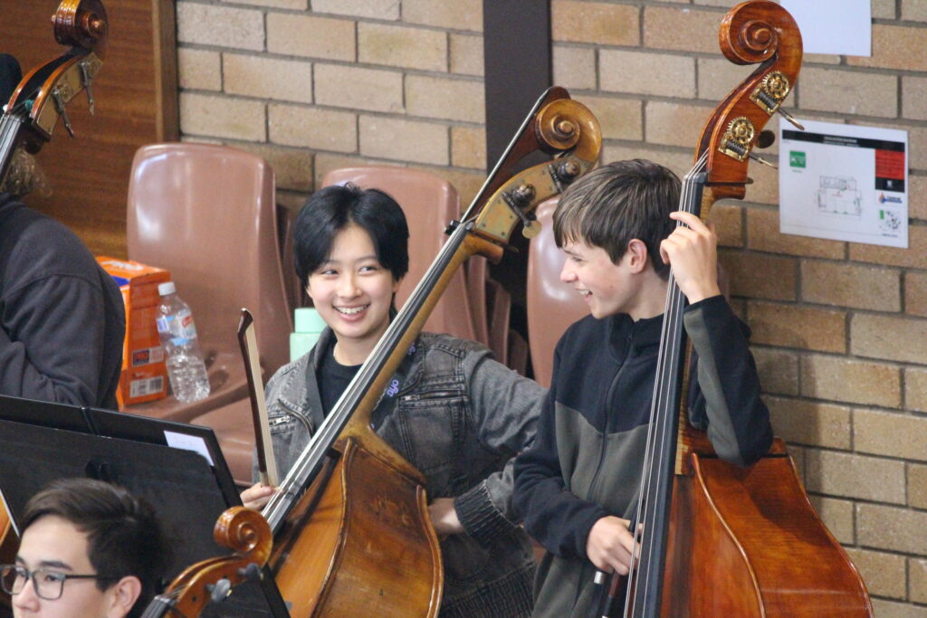 Two young double bass players laugh together during a rehearsal.