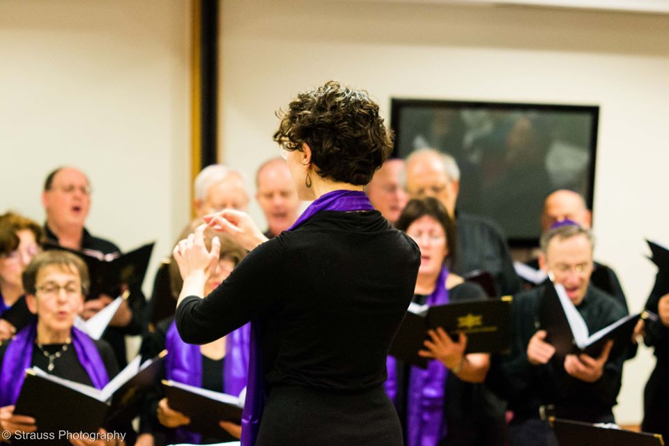 A woman conducts a choir wearing black clothes with purple sashes.