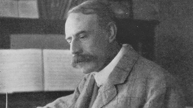 A greyscale portrait of composer Edward Elgar writing at his desk. He is wearing a suit jacket.