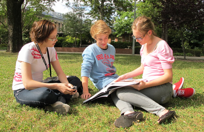 Three young musicians look at sheet music while sitting on the lawn on a sunny day.