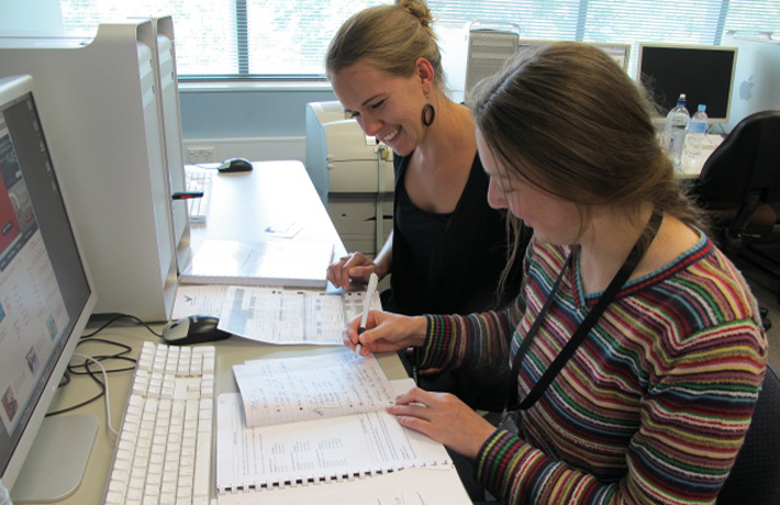 Two young women look over notes while sitting in front of a computer