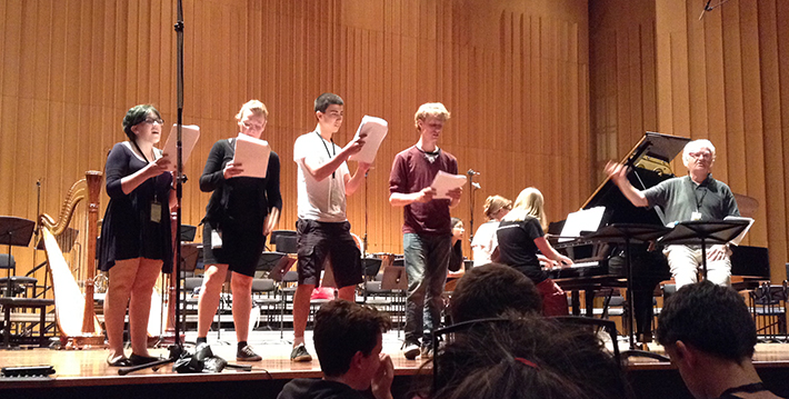 Four young musicians stand on stage, singing from sheet music. Music educator and conductor Richard Gill is seated beside them.