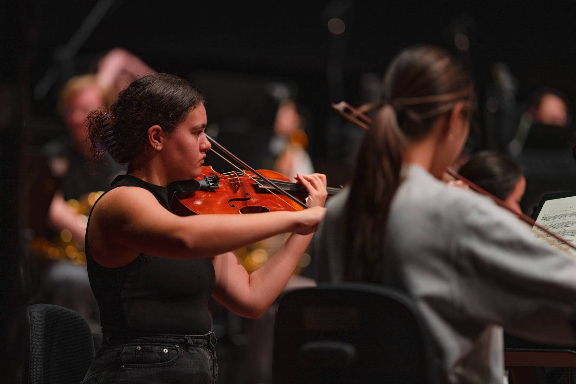 A violist of the Australian Youth Orchestra during rehearsal in the Perth Concert Hall. She is wearing a sleeveless black top and has her hair in a bun. Credit Edify Media 2023
